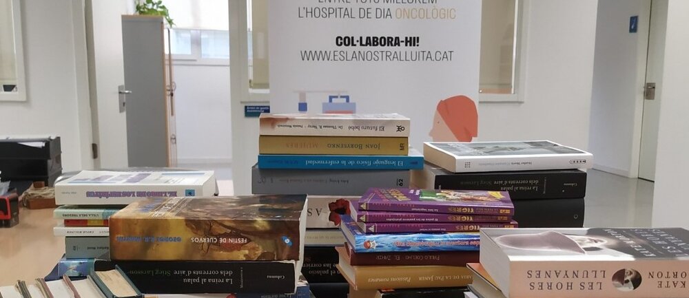 Els llibres recol·lectats apilats damunt una taula, al fons, la imatge de la campanya de mecenatge